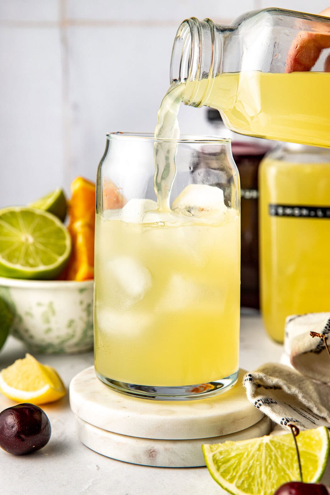 Close up view of a hand pouring a yellow Homemade Gatorade mixture into a glass of ice with fresh fruit in the background. 