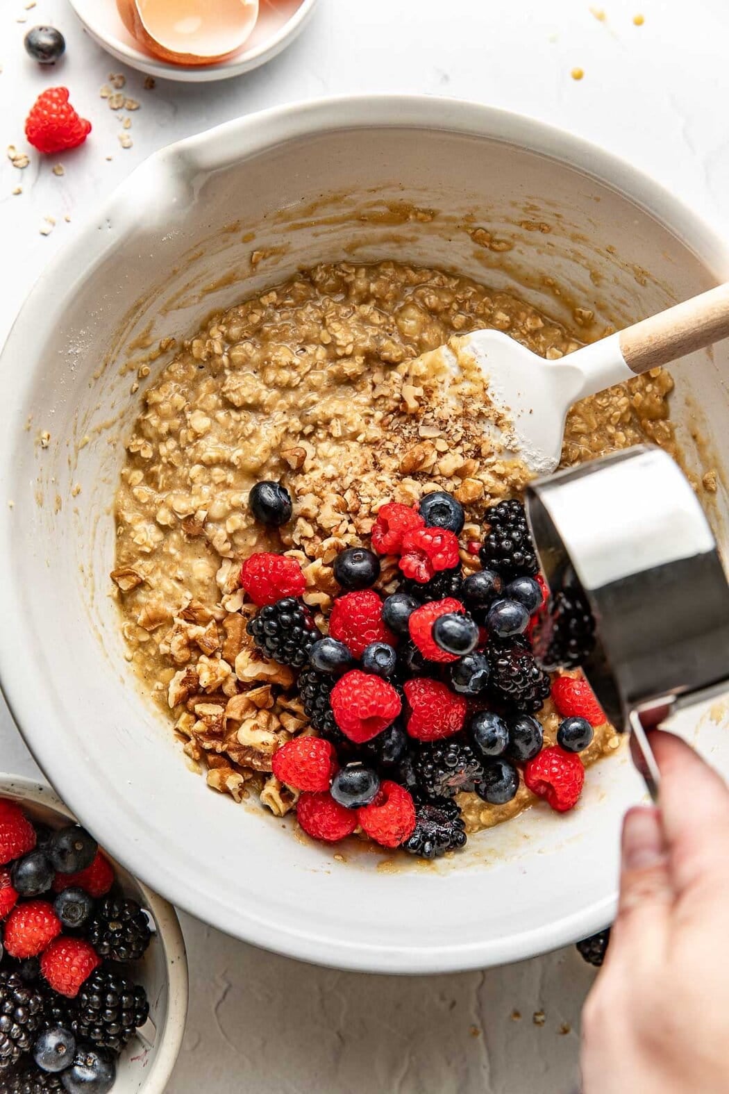 Overhead view of a bowl of an oat mixture with a cup of fresh berries being poured into the mixture. 