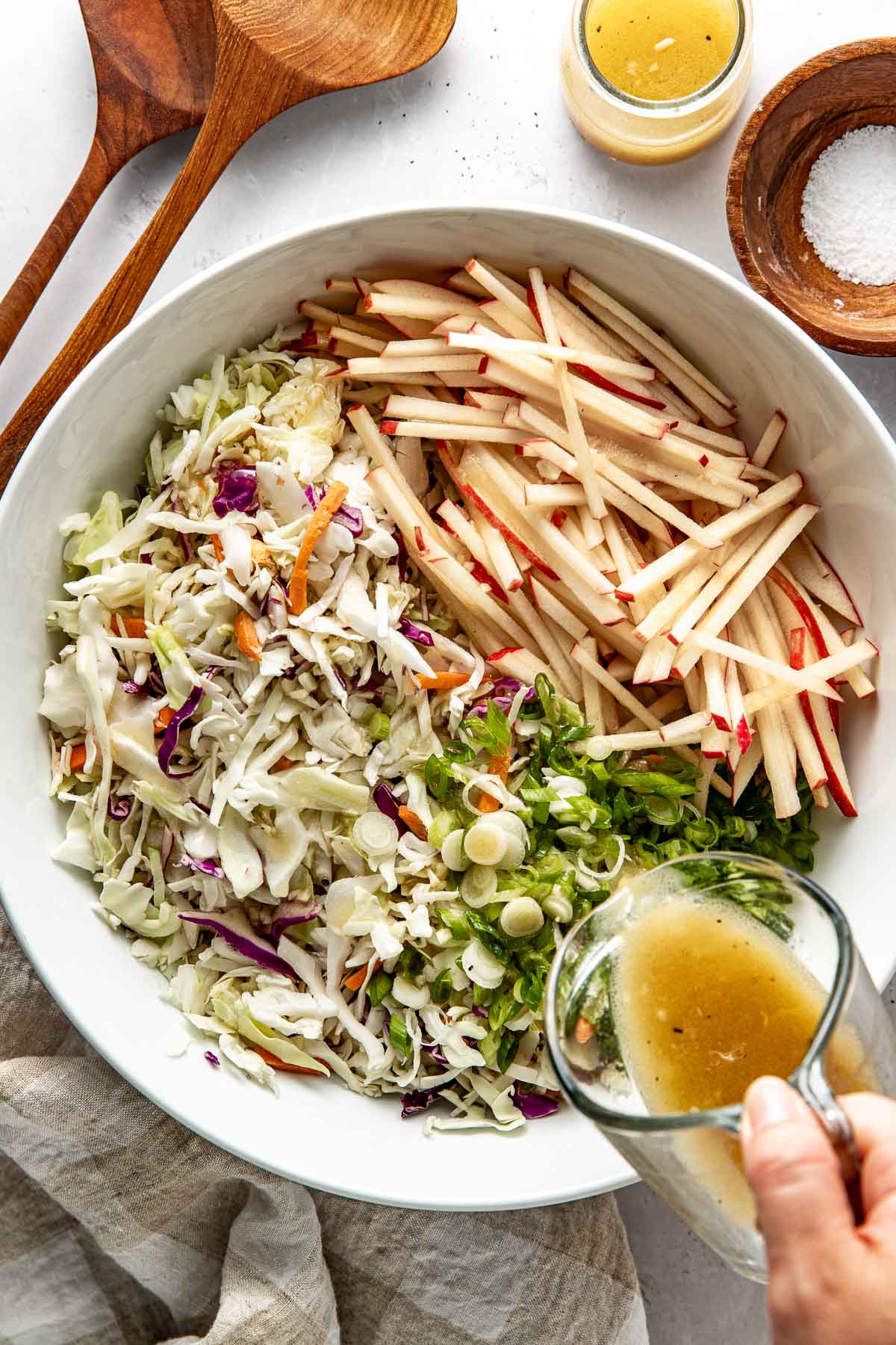 Overhead view of a white bowl filled with Apple Slaw ingredients and a hand pouring liquid dressing over the mixture. 