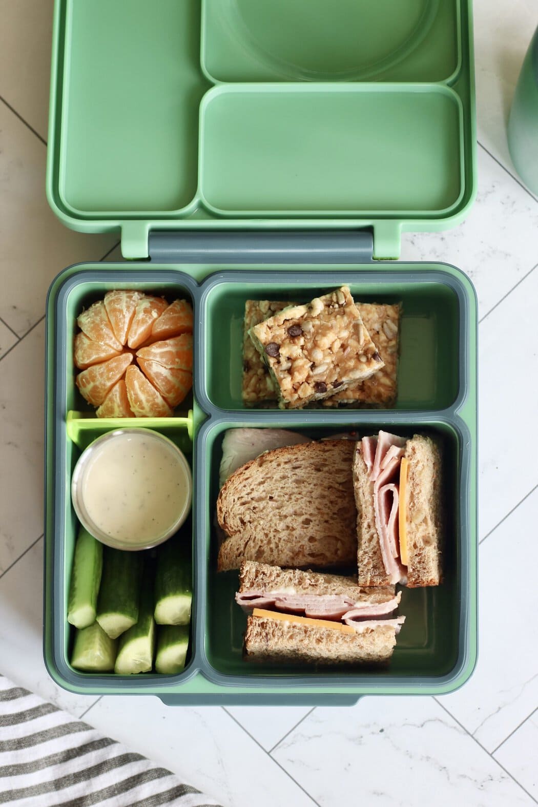 Overhead view of an open lunchbox filled with a peeled orange, cucumber sticks, homemade clif bars, and a ham and cheese sandwich. 
