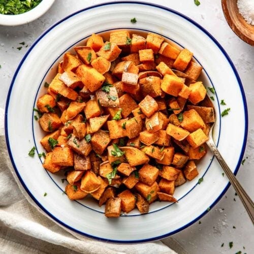 Overhead view of a white bowl of freshly Roasted Sweet Potatoes topped with fresh herbs and sprinkled with seasonings.