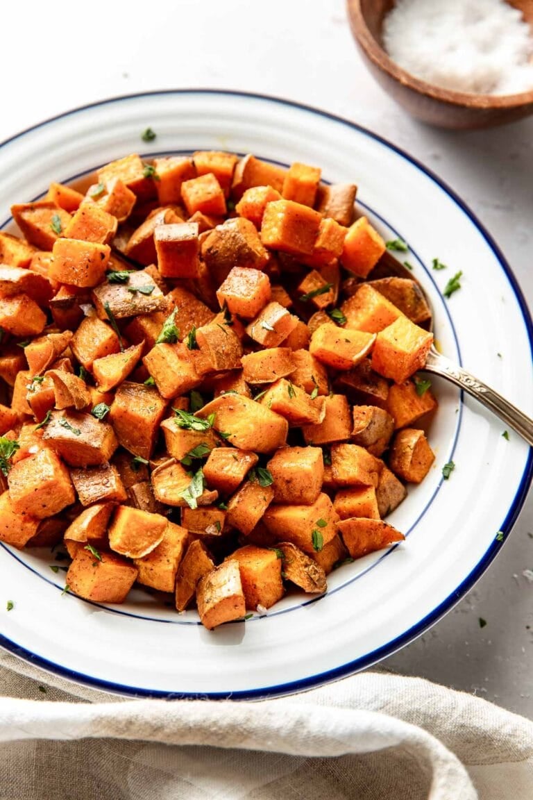 Overhead view of a white bowl filled with cubed roasted sweet potatoes topped with fresh herbs and black pepper.