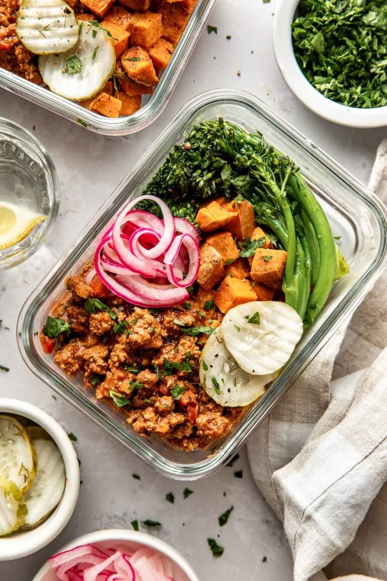 Overhead view of two glass meal prep containers filled with sloppy joe meat, roasted sweet potatoes, and broccolini topped with pickled red onions and pickle chips.