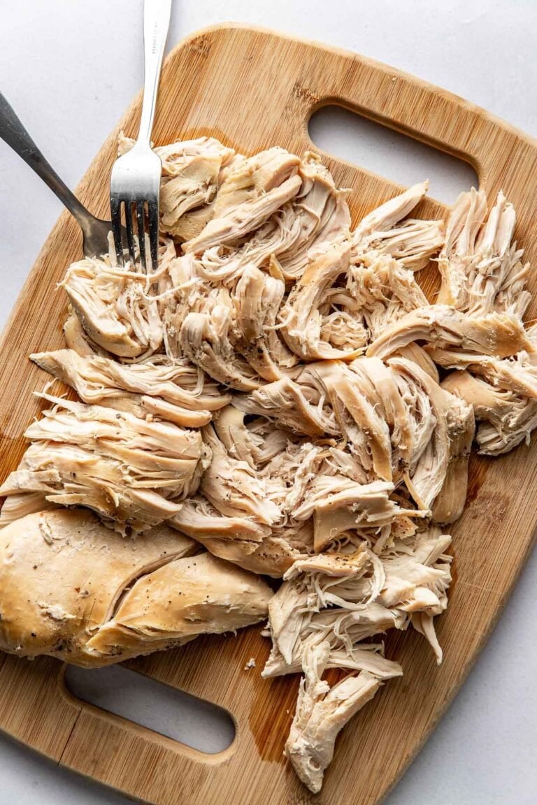 Overhead view of a wooden cutting board filled with shredded chicken next to two forks.
