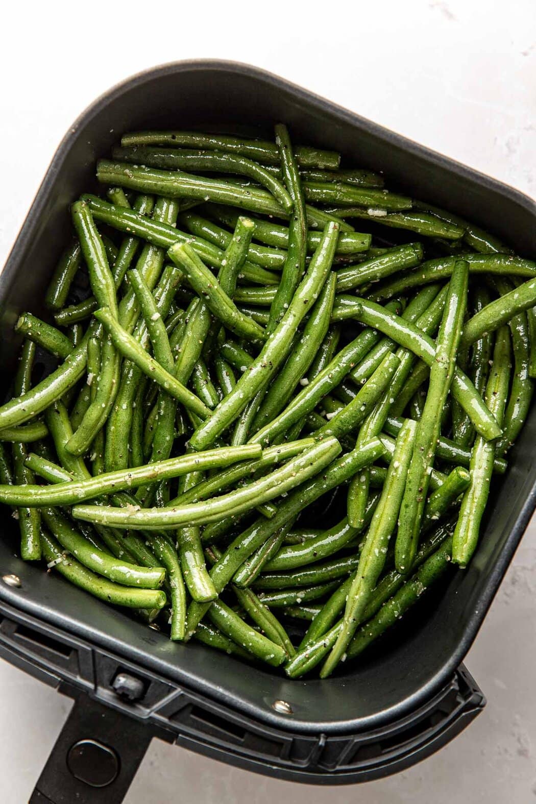 Overhead view of an air fryer basket filled with green beans tossed in avocado oil and seasoned with salt and pepper. 