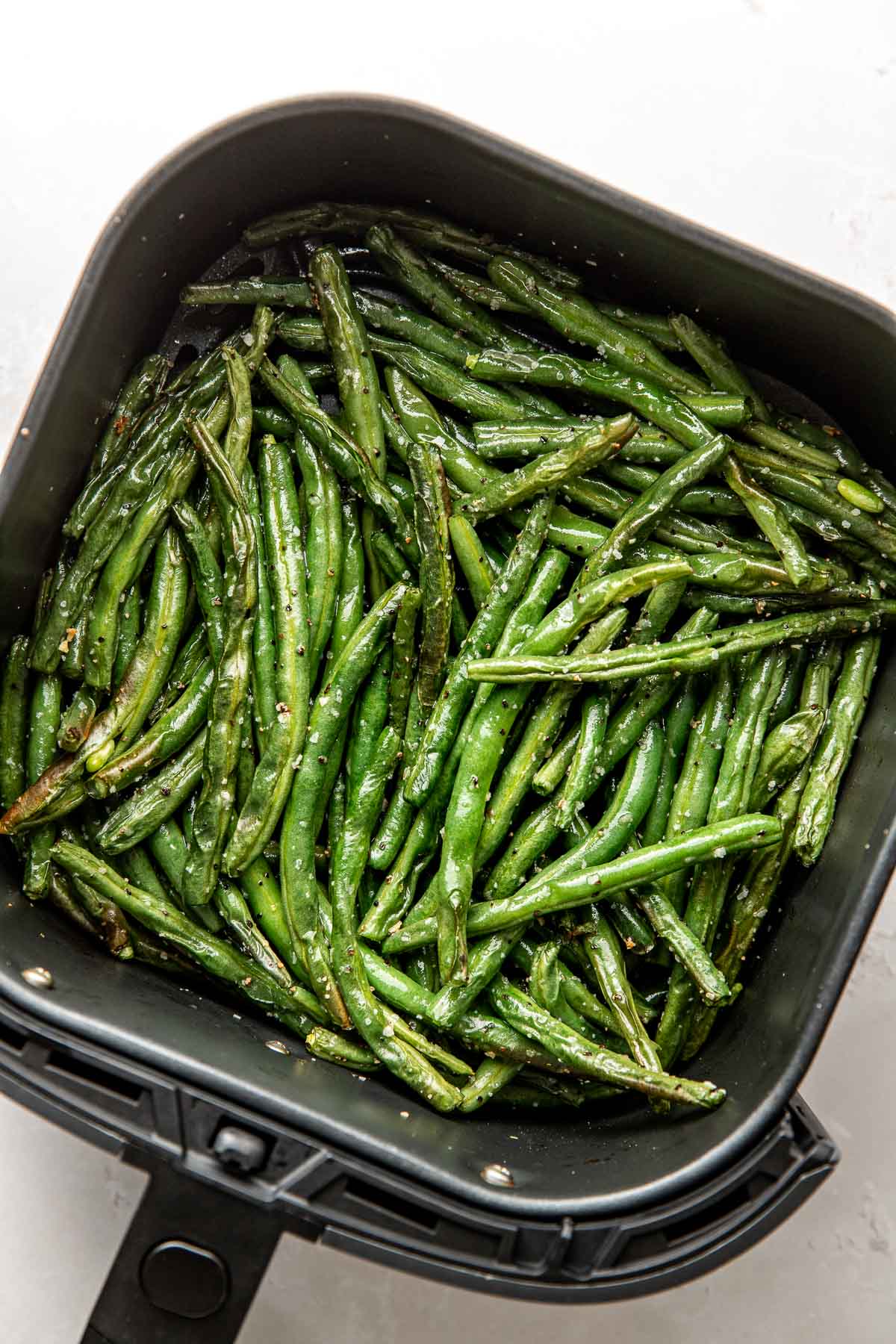 Overhead view of an air fryer basket filled with freshly roasted green beans topped with salt and pepper. 