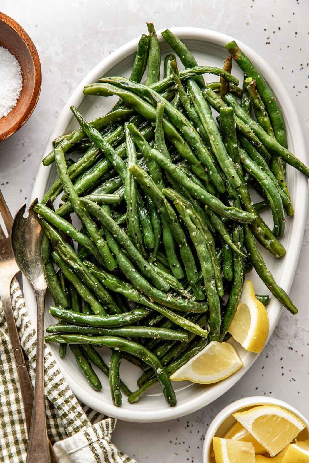 Overhead view of a white plate filled with freshly roasted green beans topped with salt and pepper and garnished with lemon wedges. 