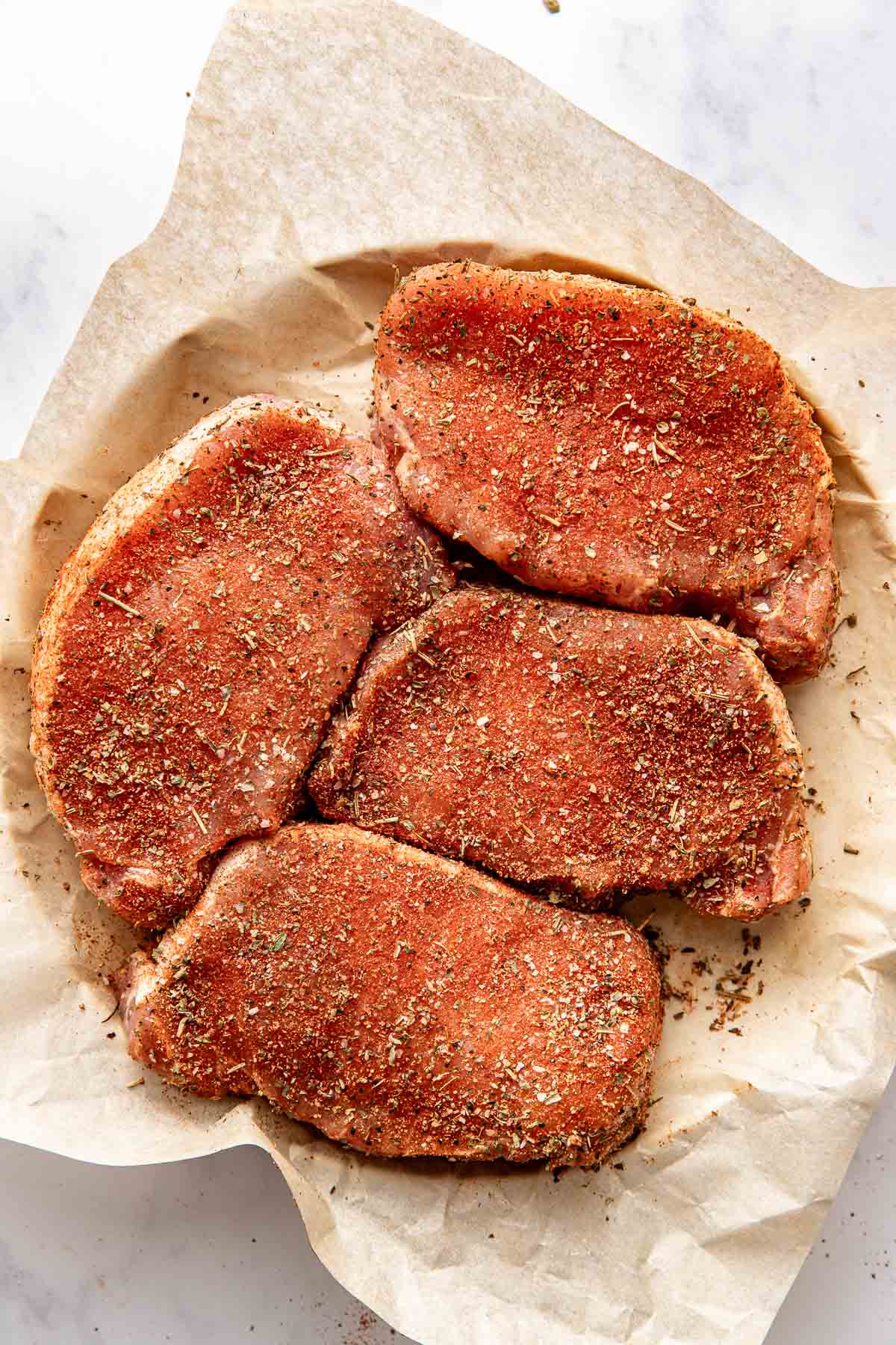 Overhead view of four perfectly seasoned pork chops ready for cooking laying on parchment paper. 