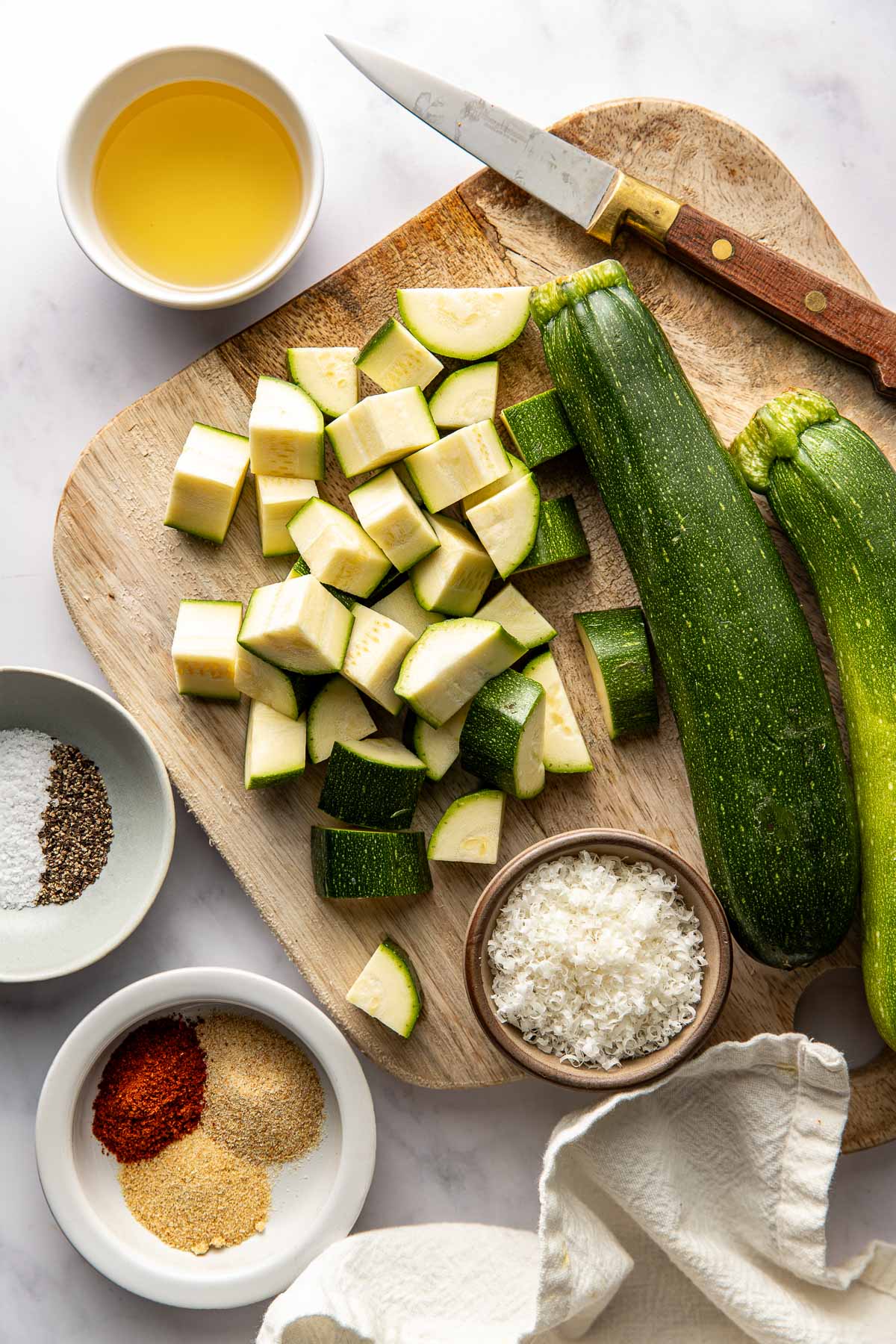Overhead view of a wooden cutting board filled with zucchini pieces surrounded by seasonings and grated parmesan cheese. 