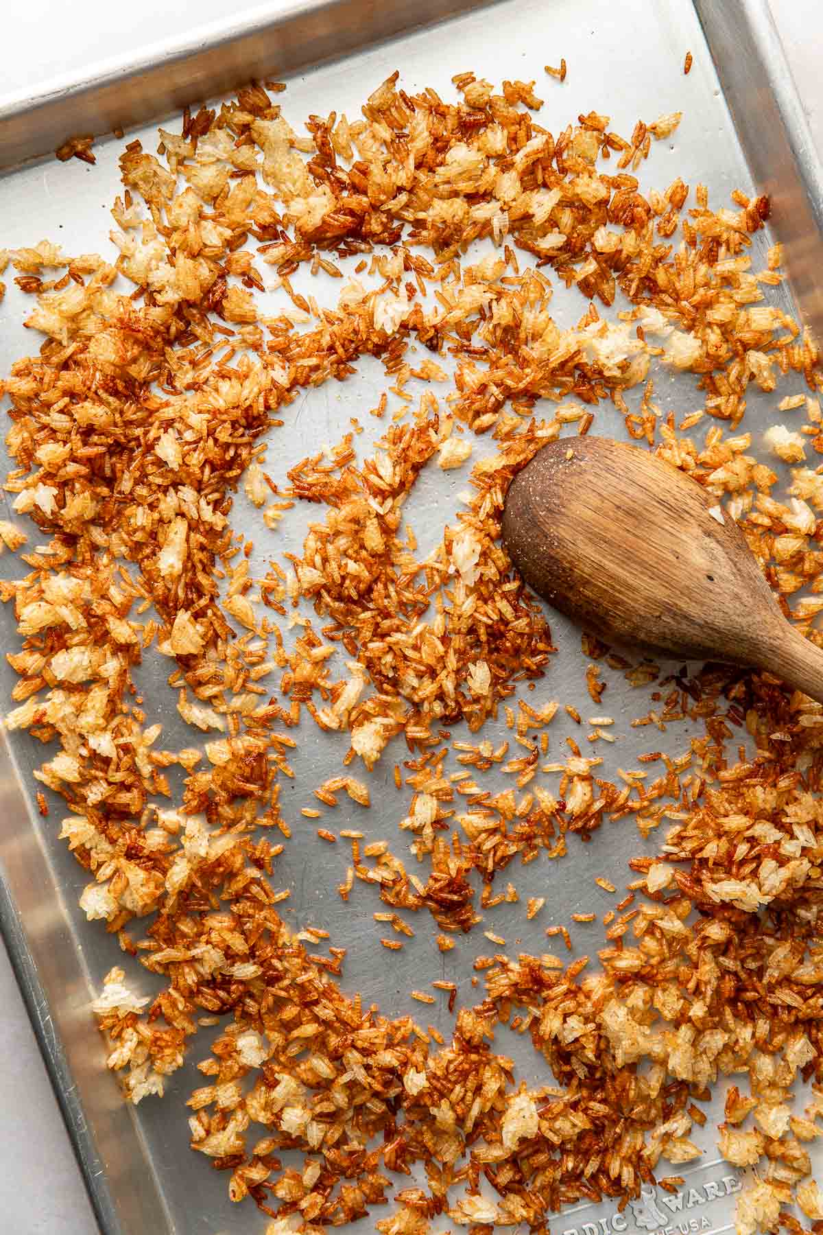 Overhead view of a sheet pan filled with freshly roasted crispy rice spread across the pan with a wooden spoon. 