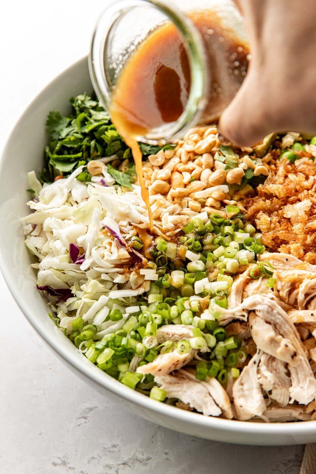 Close up view of a hand pouring a glass jar of dressing over a bowl of colorful vegetables and shredded chicken. 