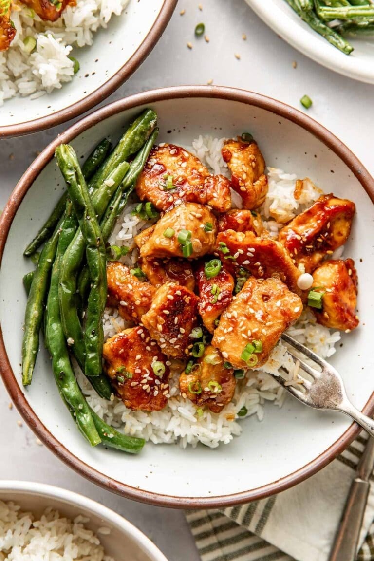 Overhead view of a plate of sesame chicken over white rice next to green beans topped with green onions and sesame seeds. 