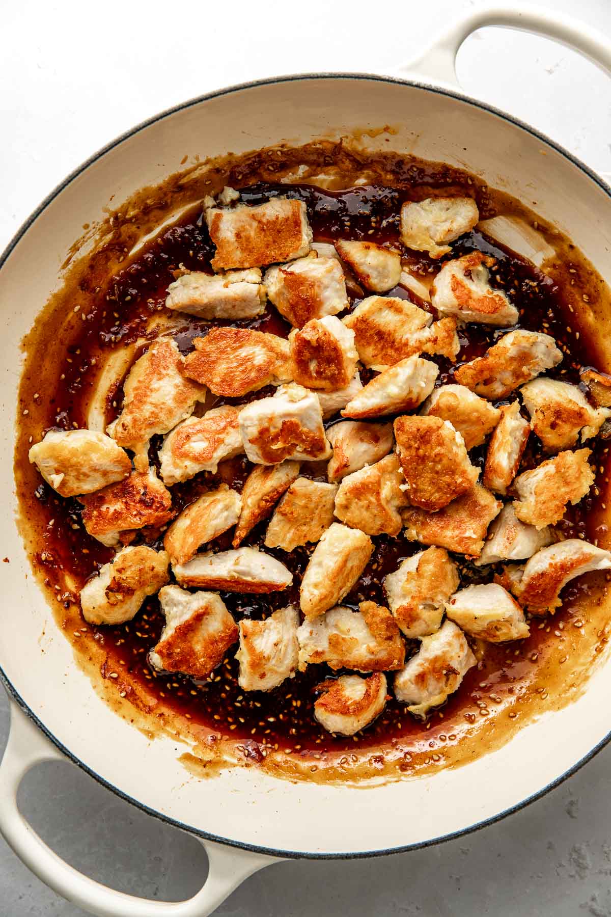 Overhead view of a skillet filled with a sesame sauce with pieces of browned chicken on top ready for stirring. 