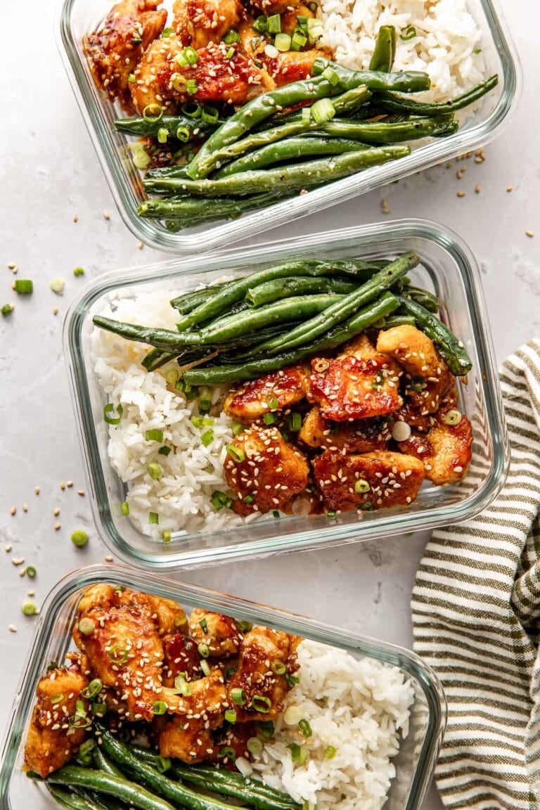Overhead view of three glass meal prep containers filled with sesame chicken, white rice, and green beans sprinkled with sesame seeds.
