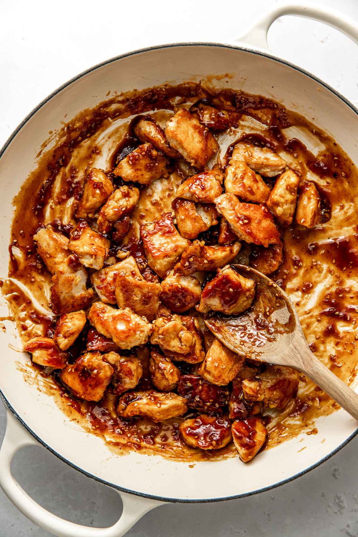 Overhead view of a skillet filled with chicken mixed in a sesame sauce with a wooden spoon. 