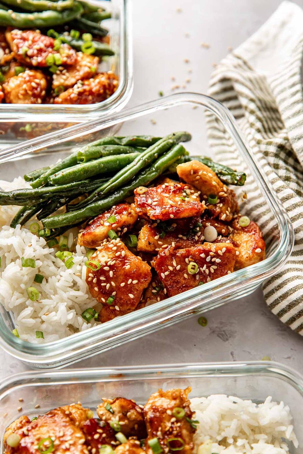 Close up view of two glass meal prep containers filled with rice, sesame chicken, and roasted green beans. 