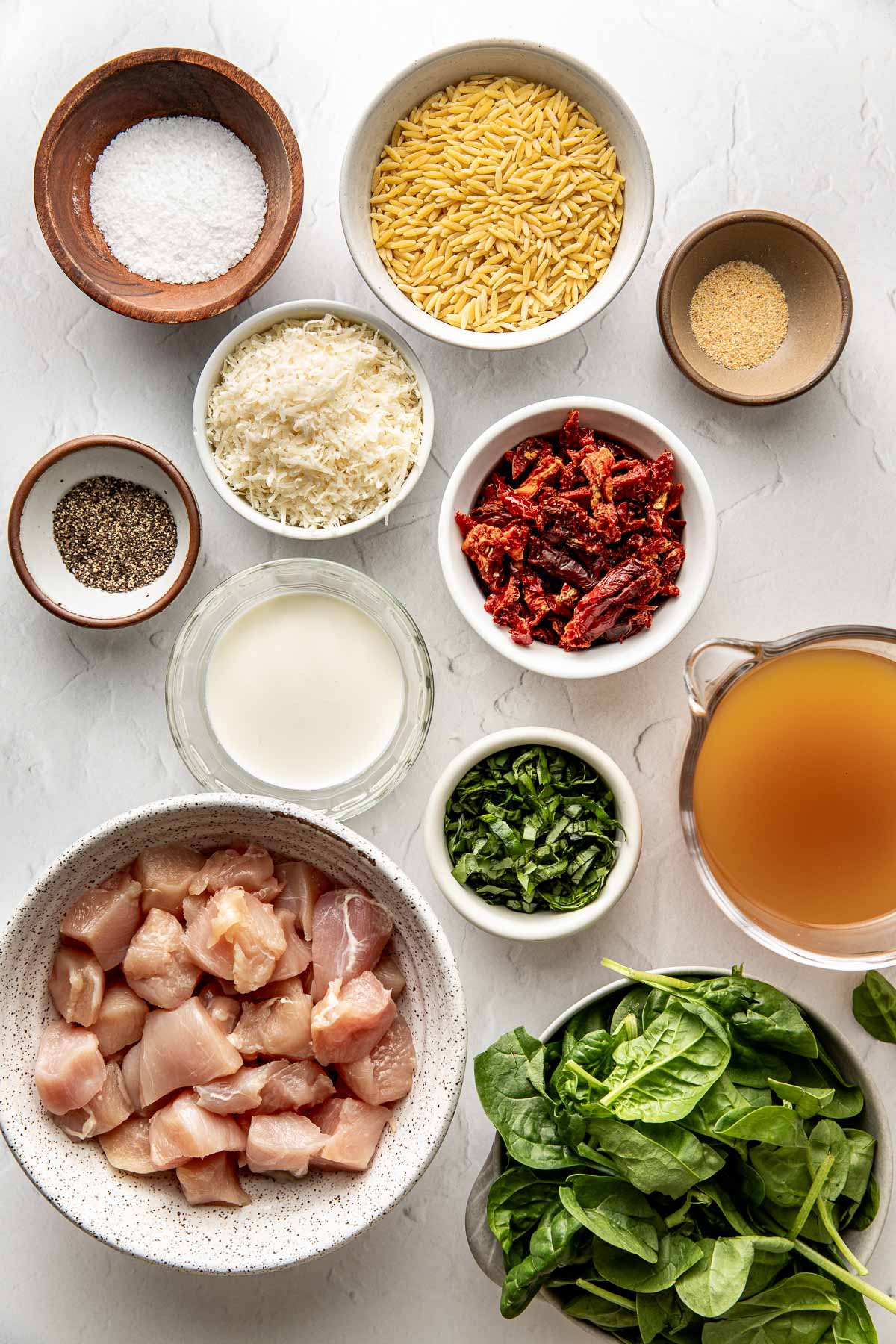 Overhead view of a variety of ingredients for Crockpot Tuscan Chicken with Orzo in different sized bowls on a white marble countertop. 
