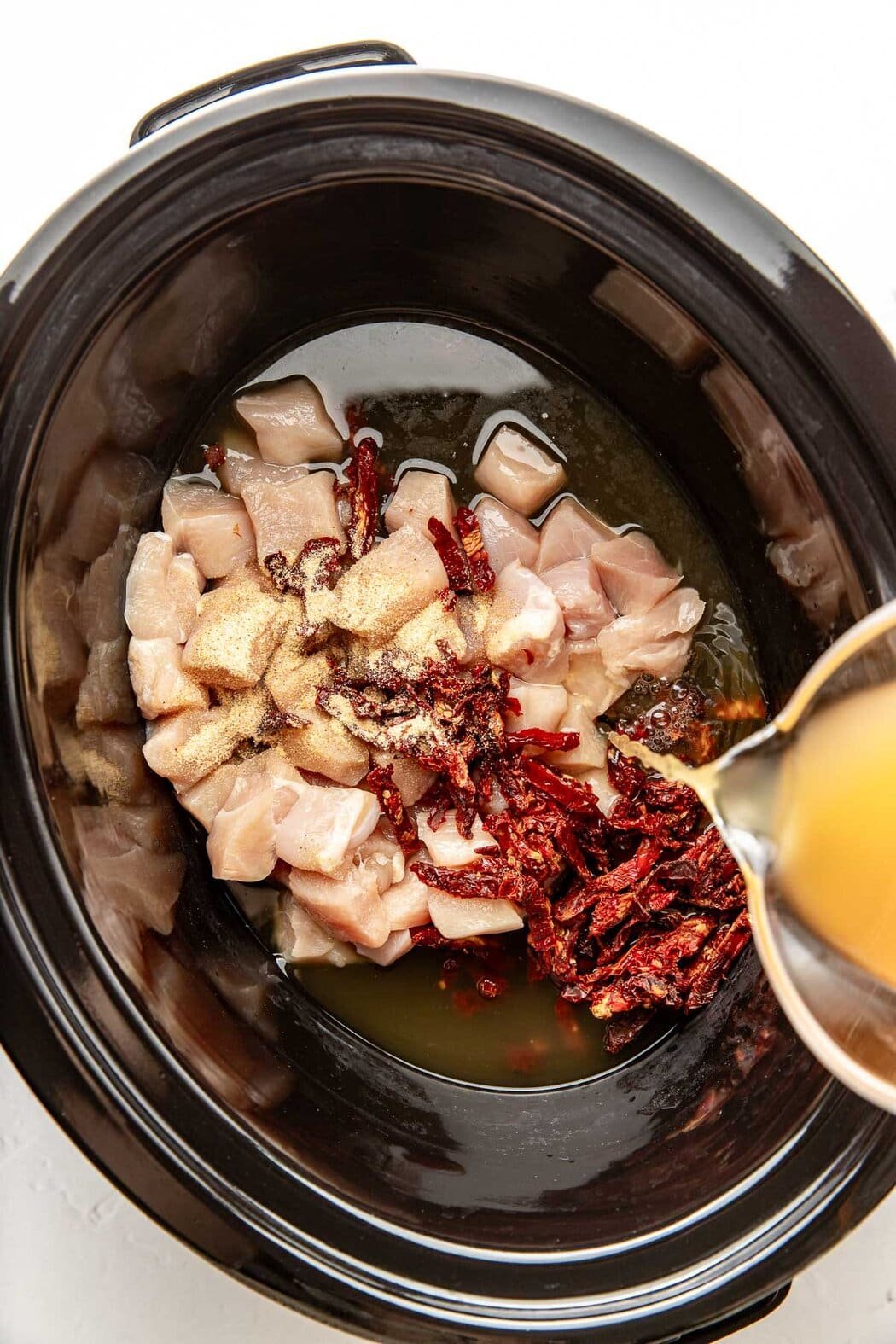 Overhead view of a slow cooker filled with raw chicken breast cubes, sun dried tomatoes, and broth being poured over them. 