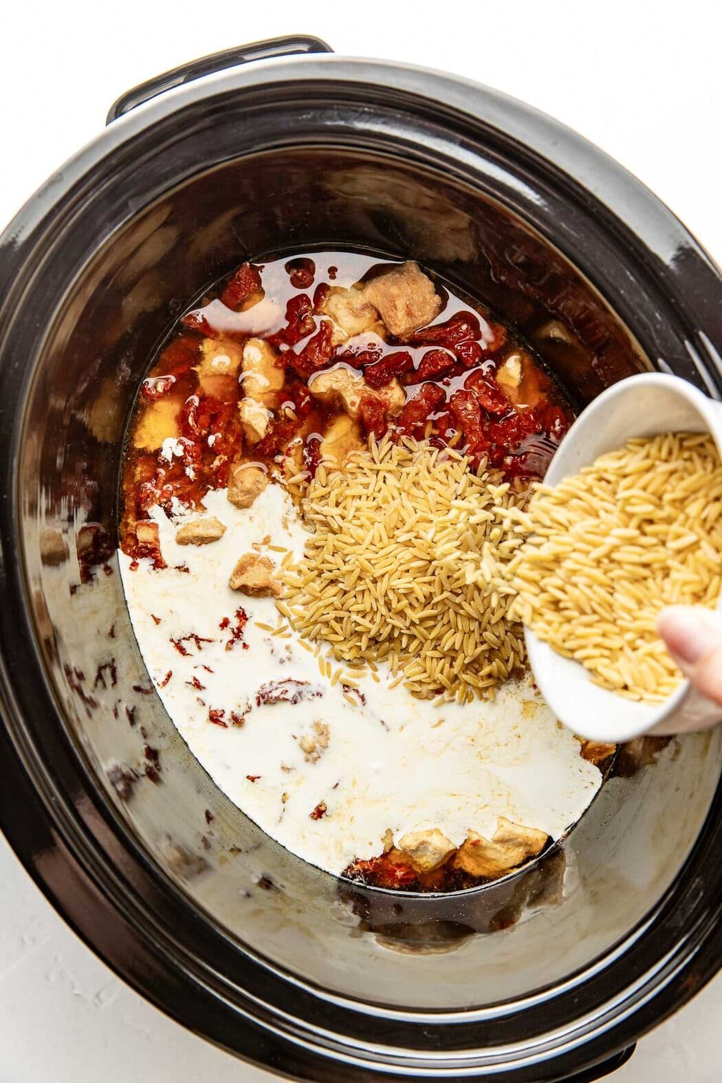 Overhead view of a slow cooker filled with chicken breast pieces, sundried tomatoes, broth, cream, and orzo being sprinkled over top. 