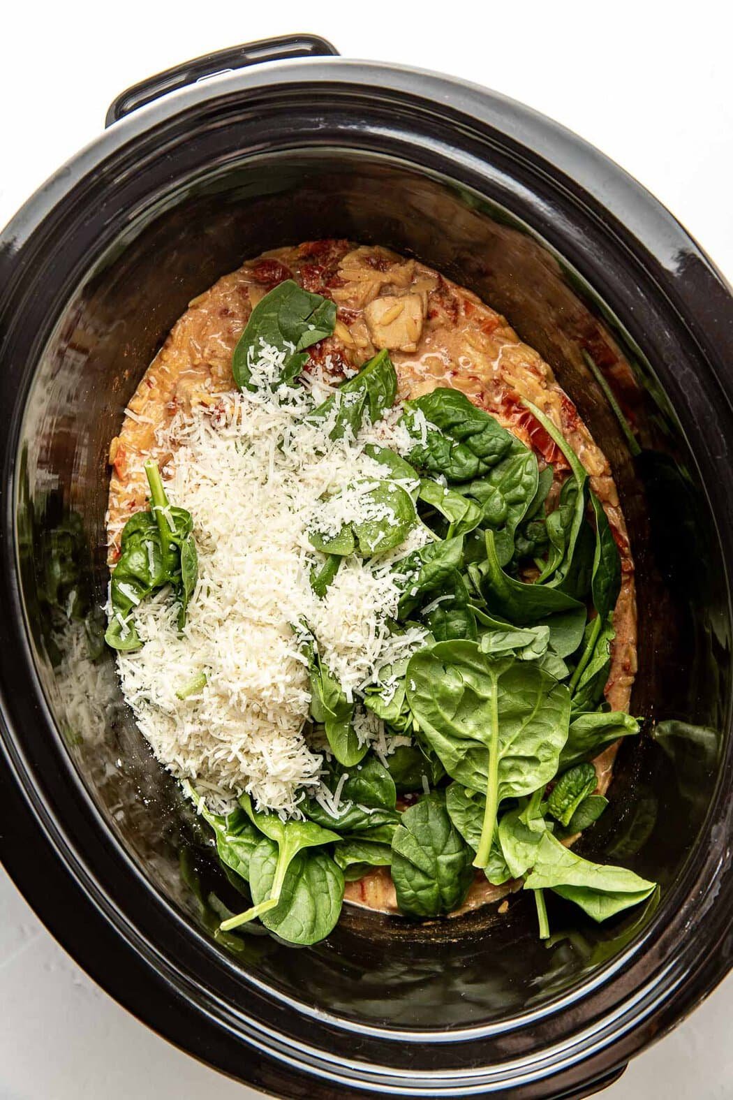Overhead view of a slow cooker filled with chicken and sun-dried tomatoes topped with fresh spinach and shredded parmesan cheese. 
