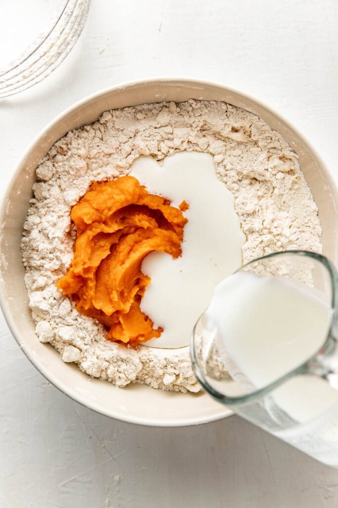 A clear glass measuring cup pouring buttermilk into a bowl with a flour mixture and mashed sweet potatoes.
