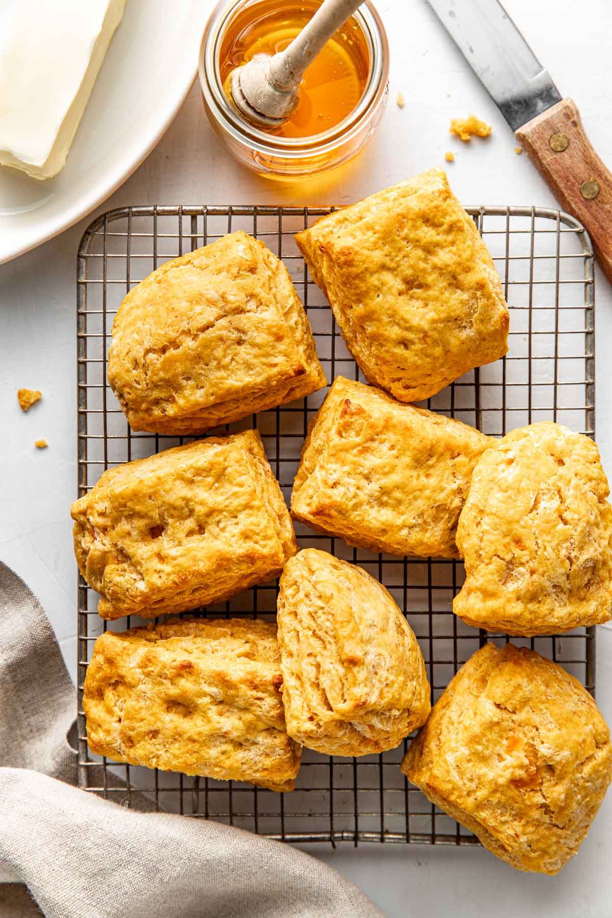 Overhead view of a batch of sweet potato biscuits on silver cooling rack with honey, butter and a knife lying next to them.