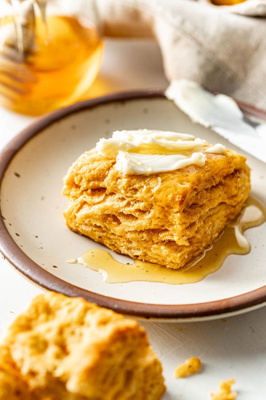 Side view view of a sweet potato biscuit slathered with soft butter and drizzled with honey on a white pottery plate with speckles and brown rim.
