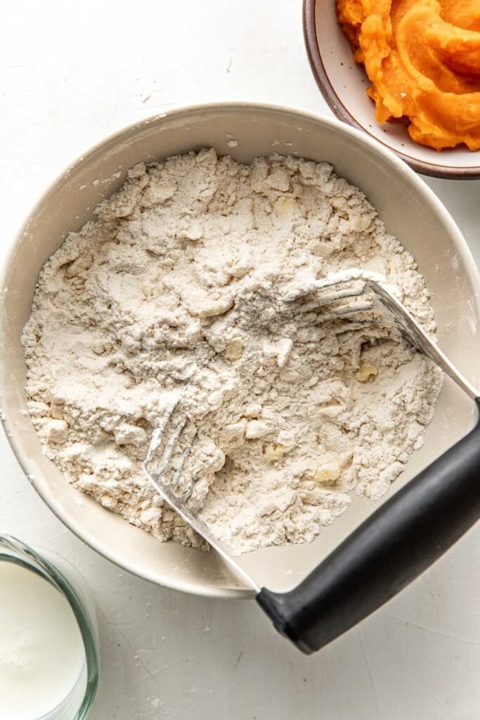 A white bowl with a flour and butter mixture and a pasty blender in the bowl in the process of making sweet potato biscuits.