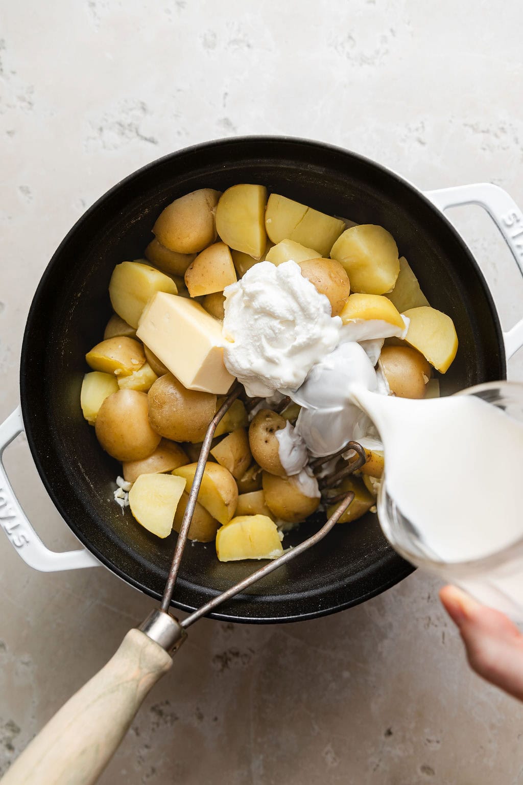 Overhead view of a hand pouring cream over freshly boiled potatoes in a Dutch oven. 