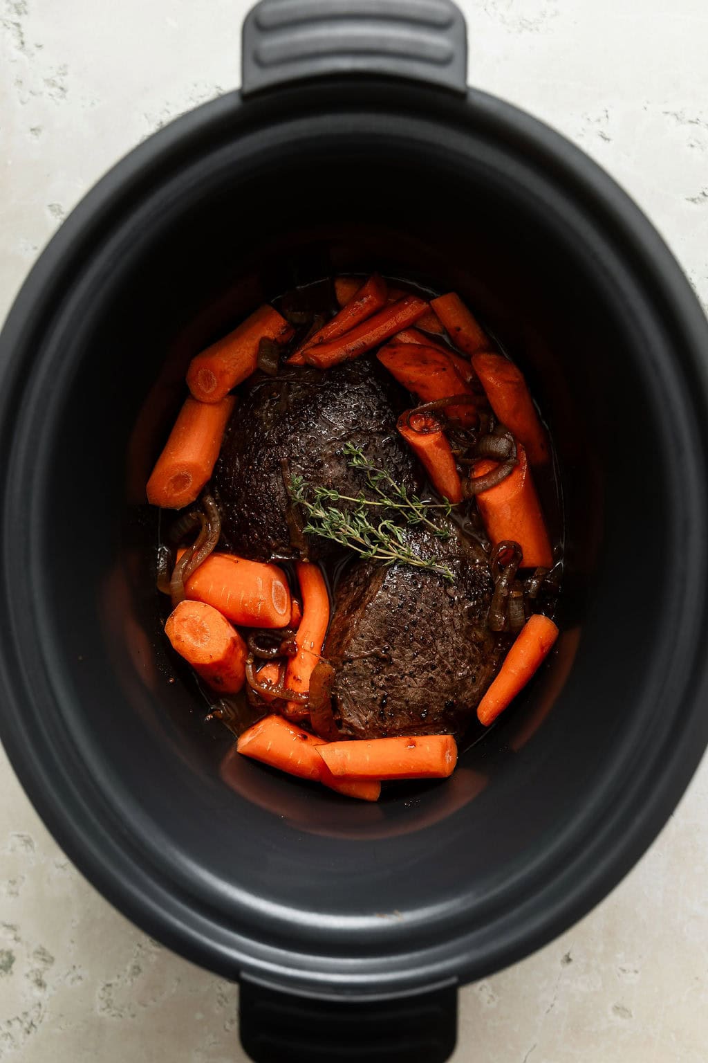 Overhead view of a slow cooker filled with venison roast with carrots around it ready for cooking. 