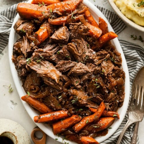 Overhead view of an oval platter filled with shredded venison meat surrounded by carrots and topped with fresh herbs.