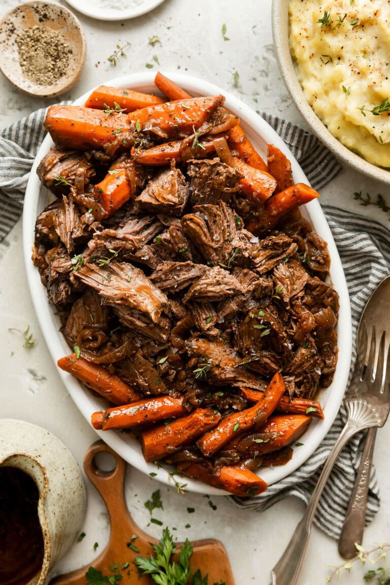 Overhead view of a platter filled with shredded venison roast surrounded by carrots and sprinkled with fresh herbs.