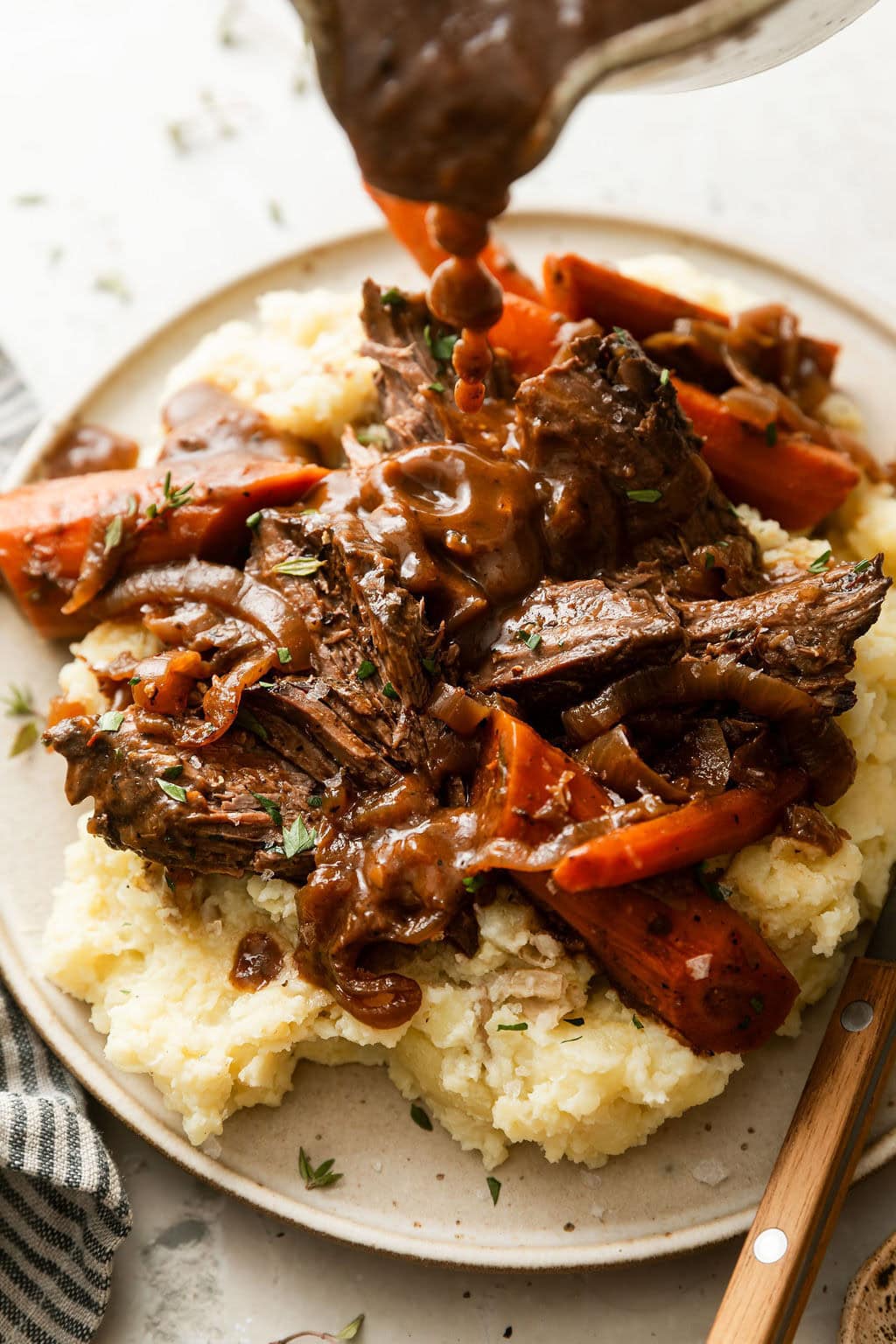 Close up view of brown gravy being poured over a plate of mashed potatoes topped with venison and carrots. 