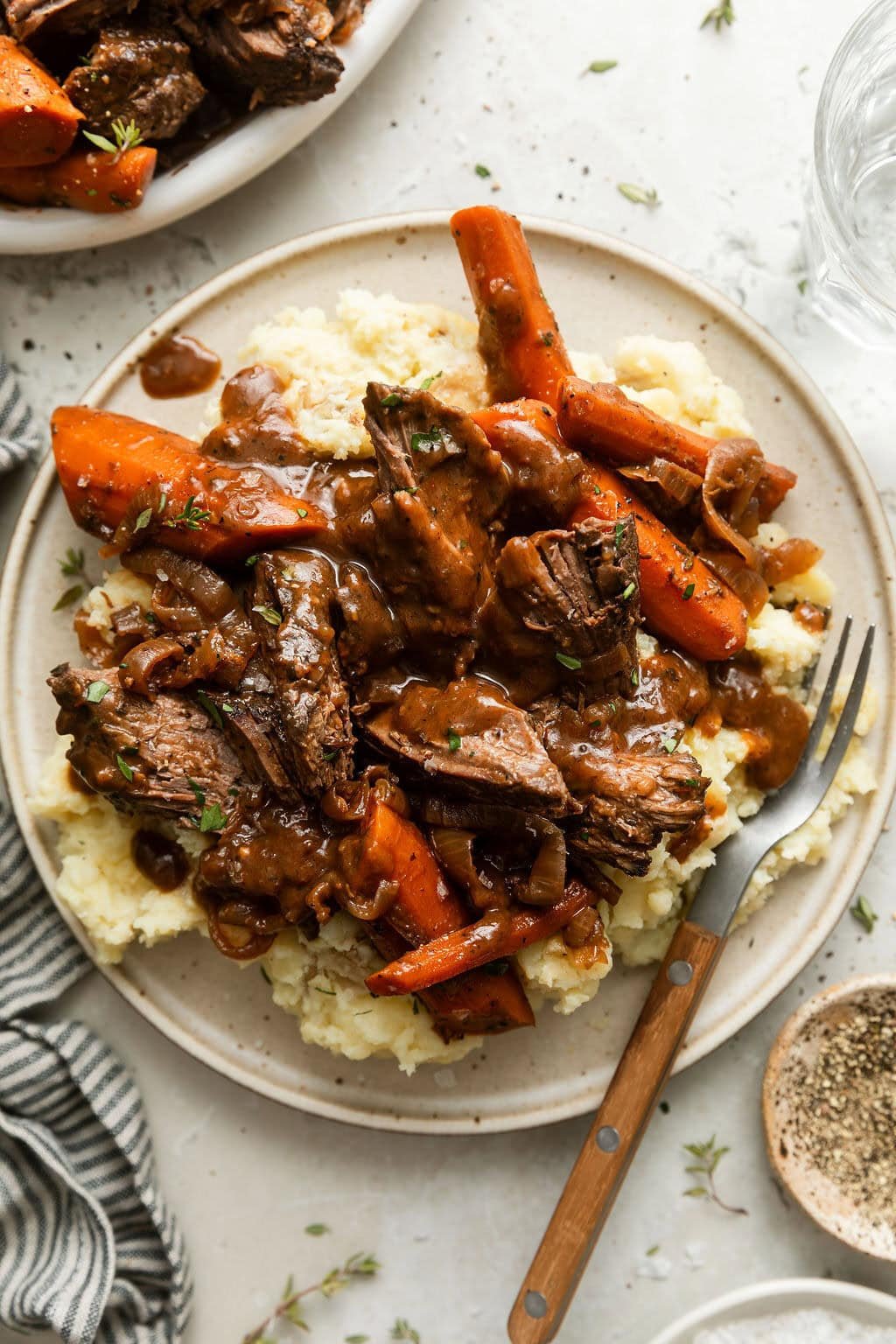 Close up view of a plate of mashed potatoes topped with shredded venison roast and carrots with gravy on top sprinkled with fresh herbs and black pepper. 
