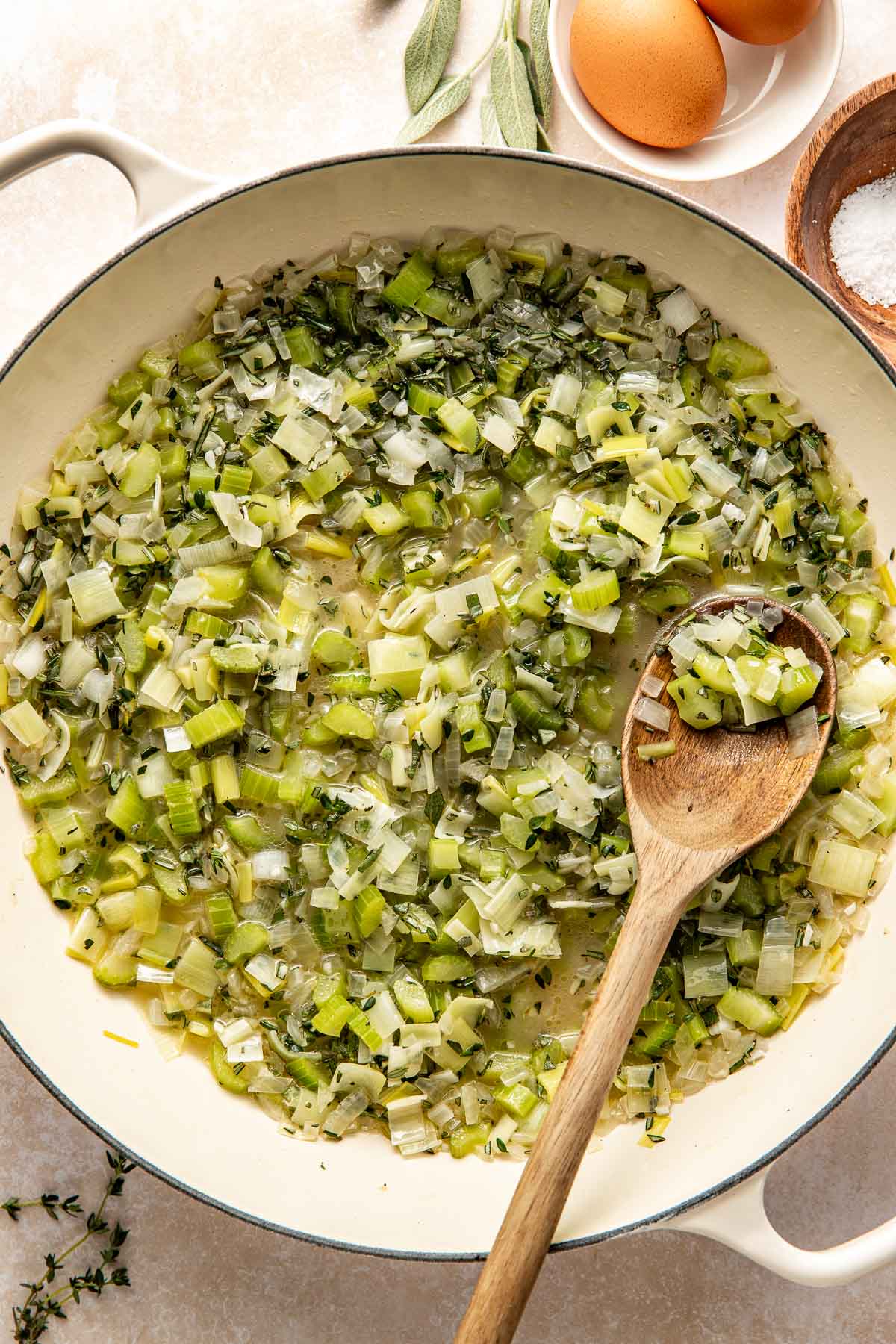 Overhead view of a white skillet filled with onions, celery, and leeks in butter sautéing.