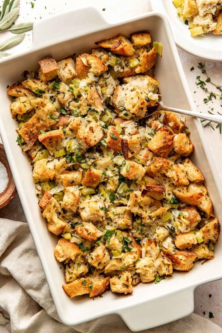Overhead view of a white baking dish filled with stuffing topped with fresh herbs and black pepper. 