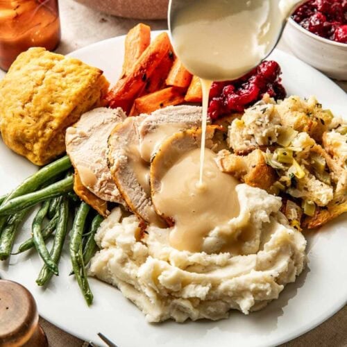 Overhead view of a white plate filled with a thanksgiving variety of foods including sliced turkey with gravy being poured over it.