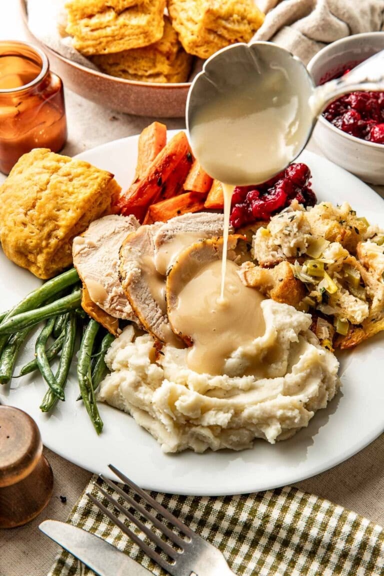 Close up view of a plate filled with thanksgiving foods and turkey gravy being poured over the turkey and mashed potatoes.