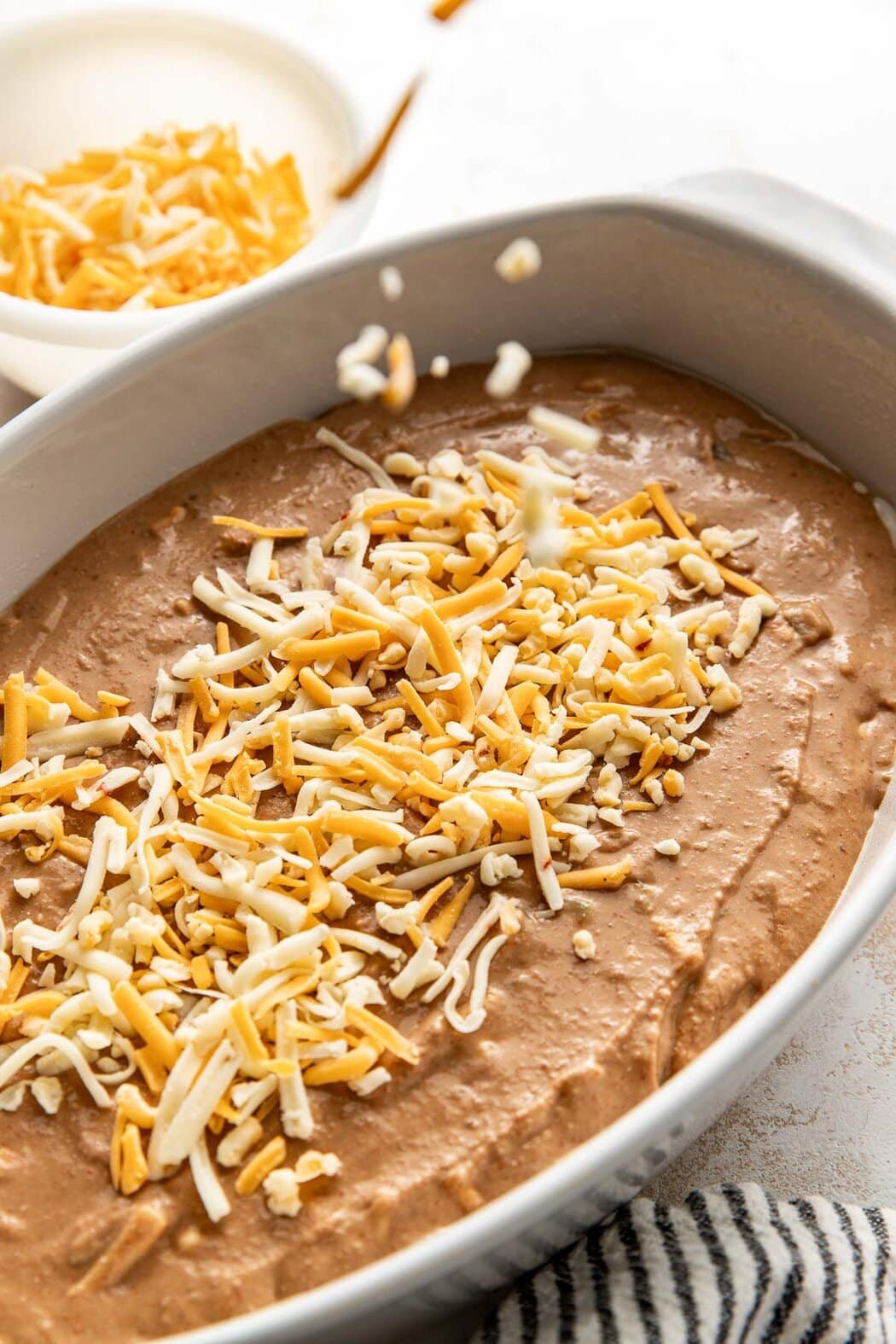 Close up view of a white oval baking dish filled with Homemade Bean Dip and topped with shredded cheese.