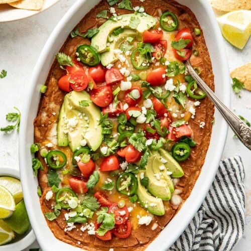 Overhead view of a white oval baking dish filled with homemade bean dip topped with fresh cilantro, diced tomatoes, and sliced jalapenos.