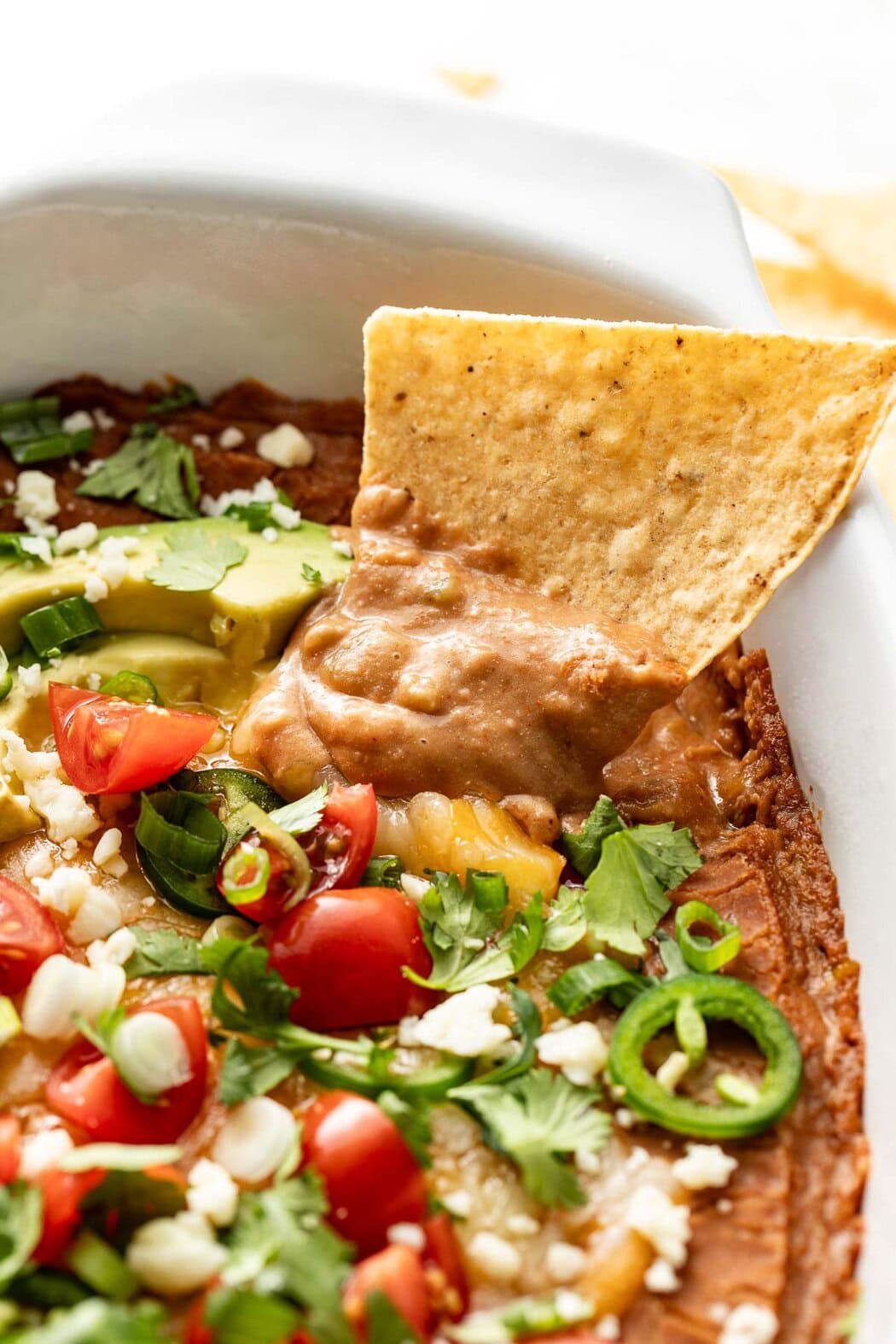 Close up view of a tortilla chip being dipped into a freshly baked Homemade Bean Dip topped with fresh cilantro, tomatoes, and sliced jalapeno.