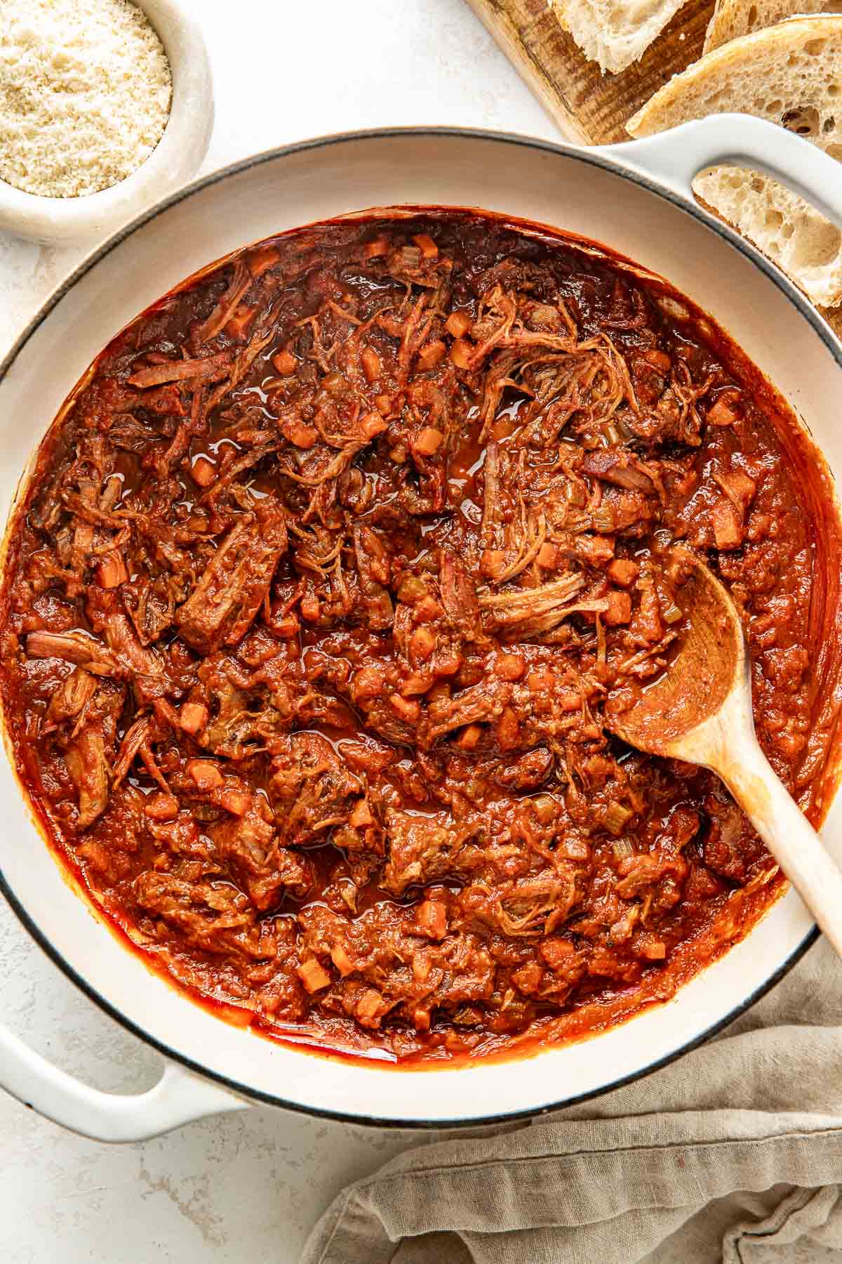 Overhead view of a white skillet filled with tomato sauce with meat for beef ragu being stirred with a wooden spoon.