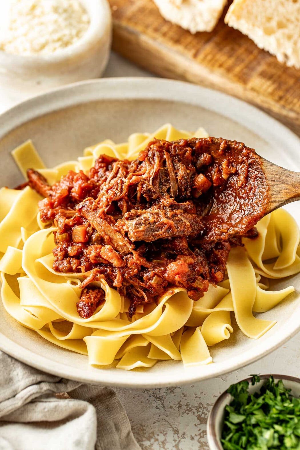 Close up view of a wooden spoon topping a plate of noodles with beef ragu.