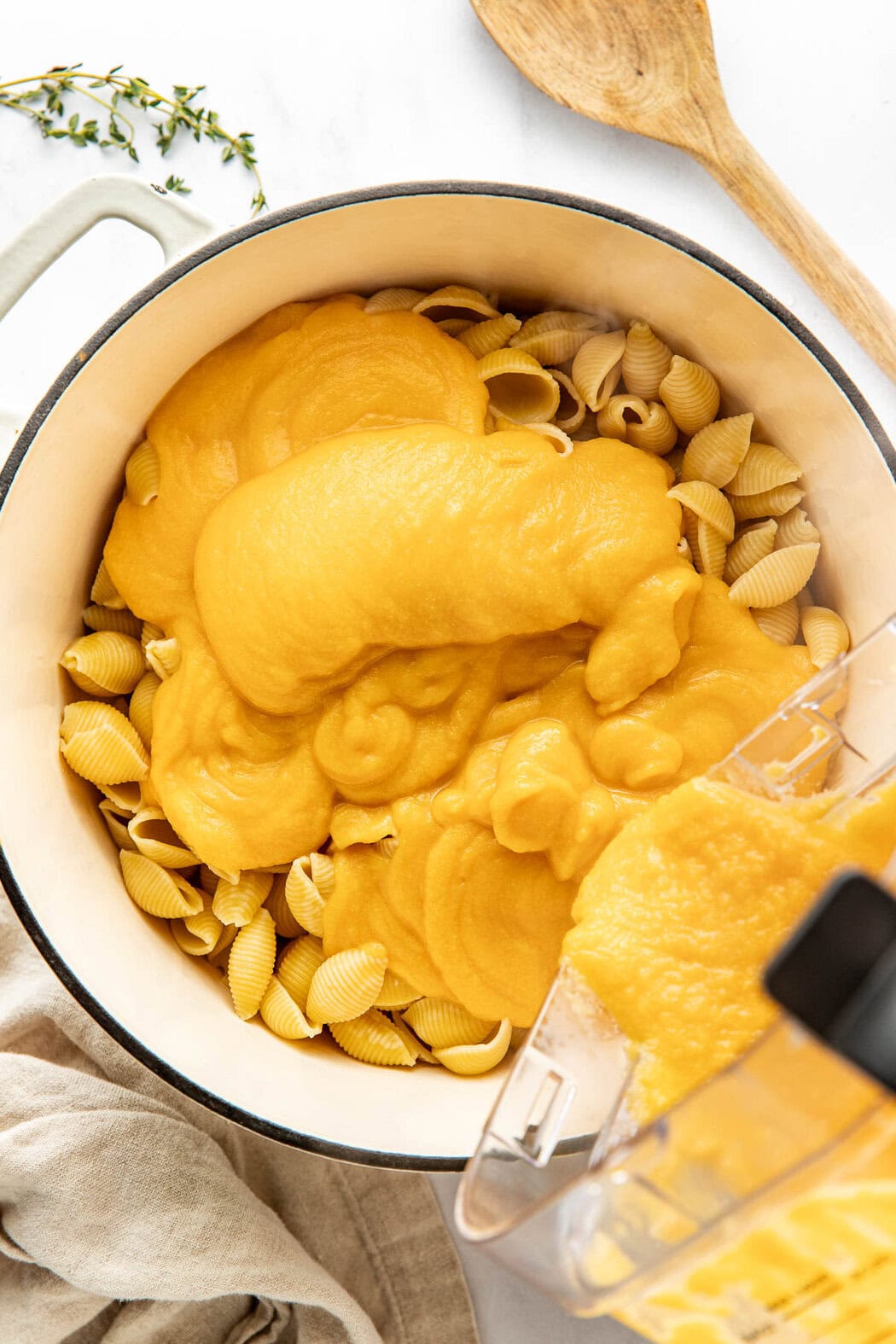 Overhead view of a pot of shell pasta noodles with a blender pouring a butternut squash sauce mixture over the top.