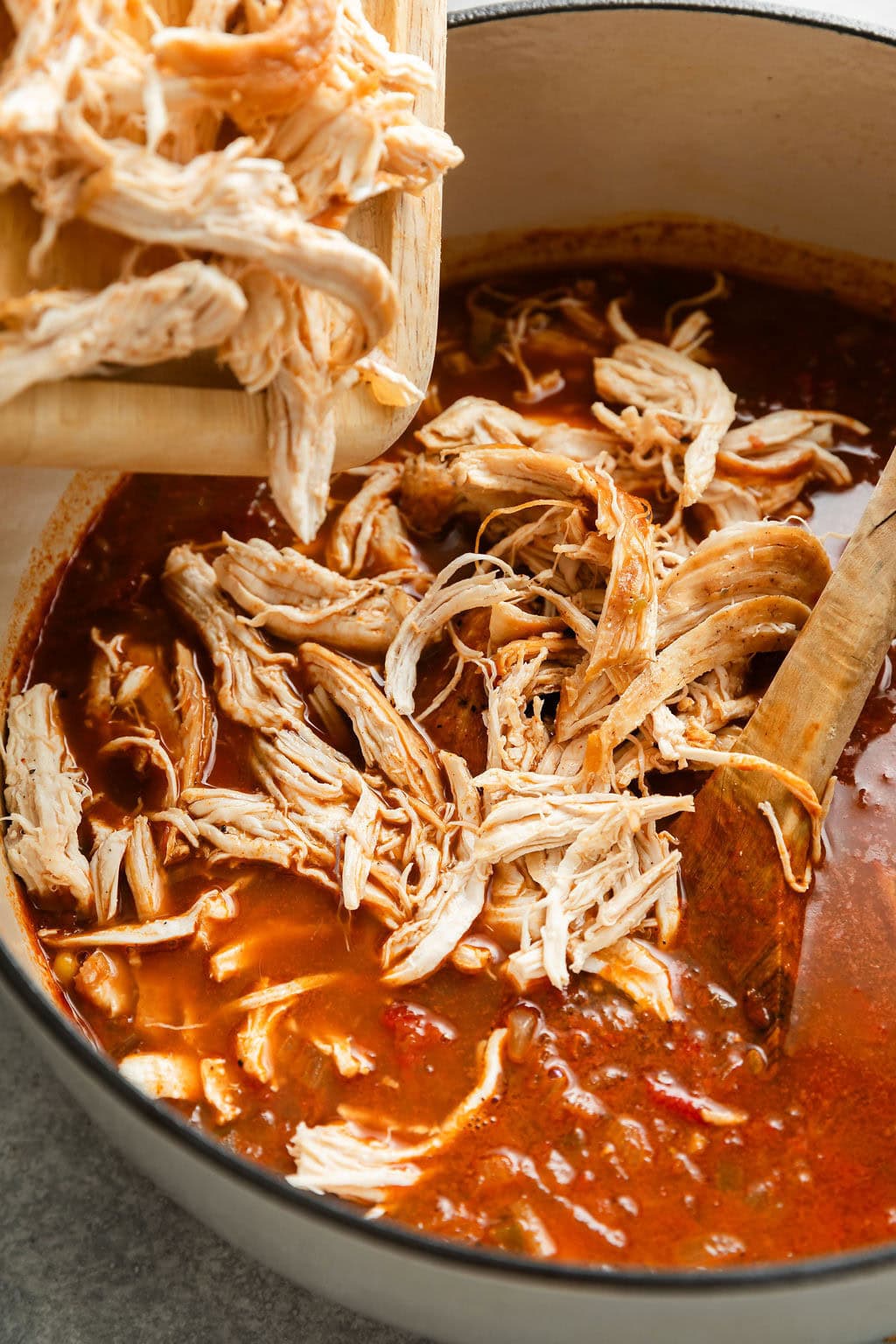 Close up view of shredded chicken going into a pot filled with a tomato broth.