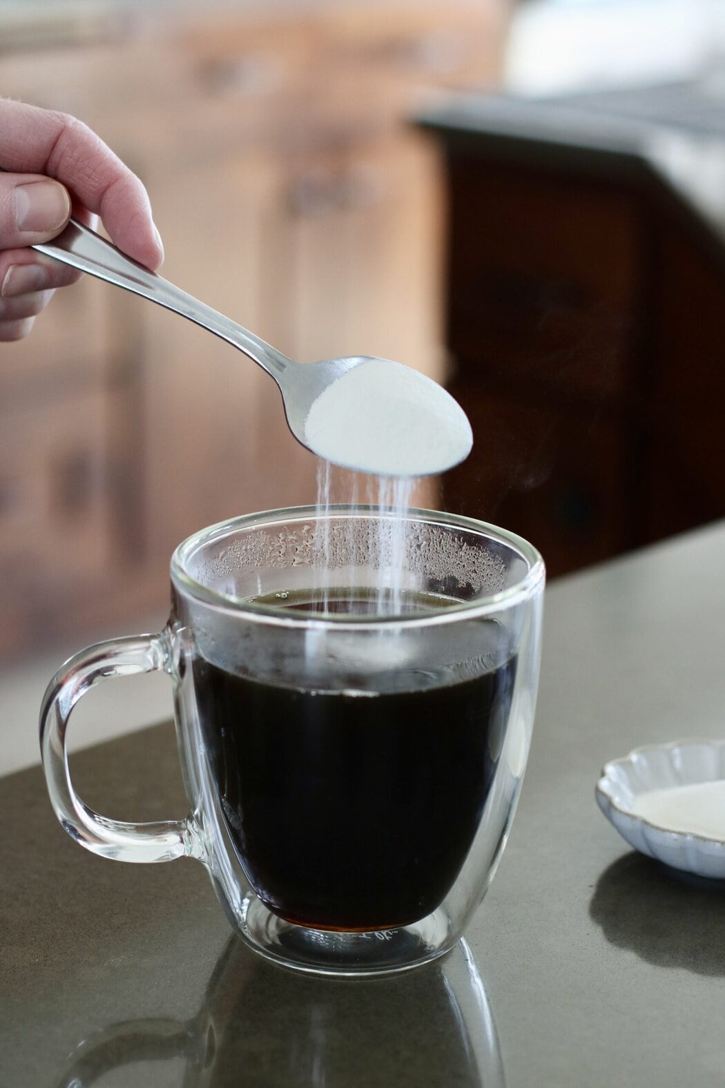 Close up view of a hand pouring a spoonful of collagen into a glass mug of coffee.