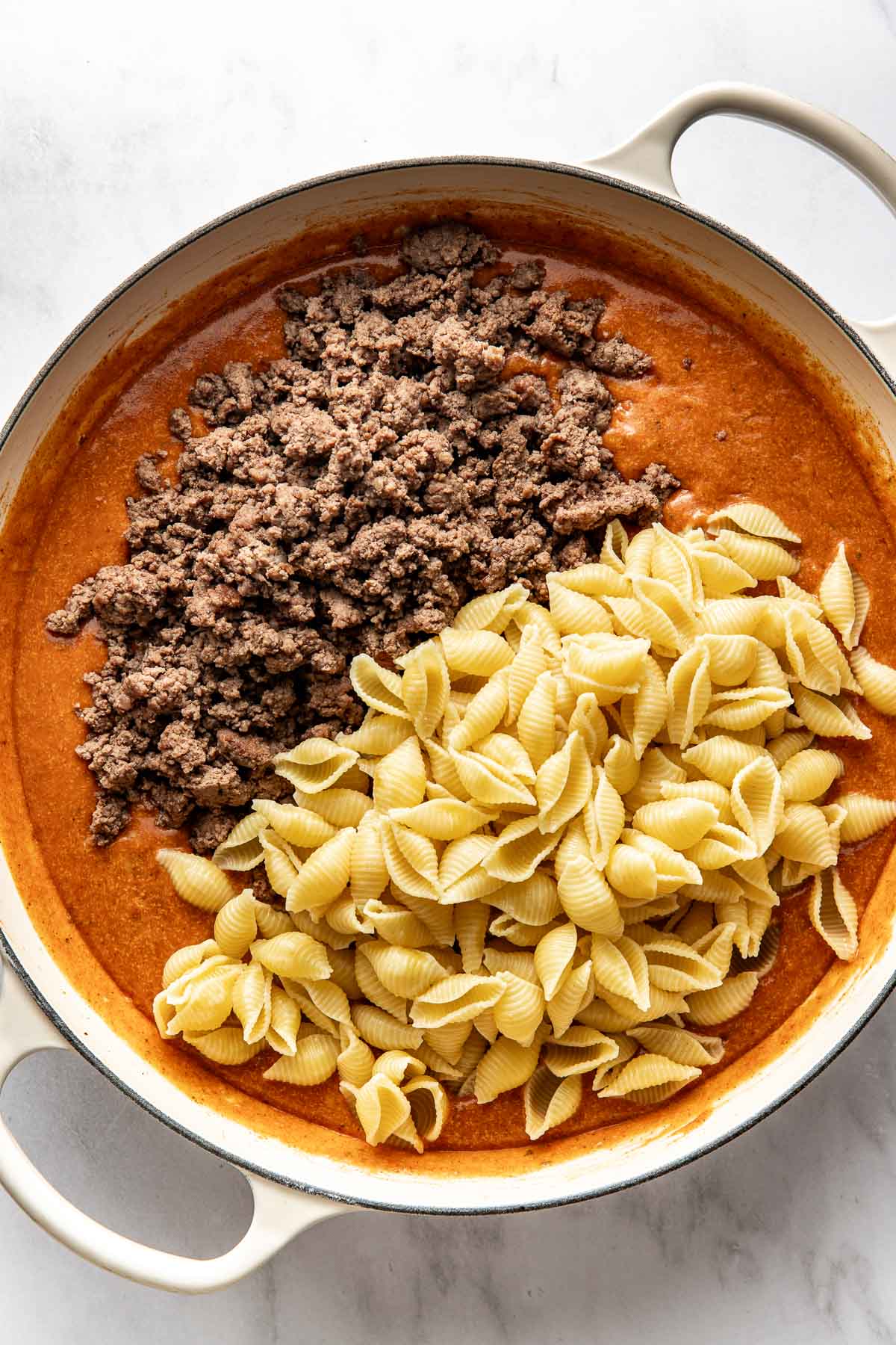 Overhead view of a white skillet filled with a tomato sauce topped with ground beef and cooked pasta ready to be stirred into the mixture.