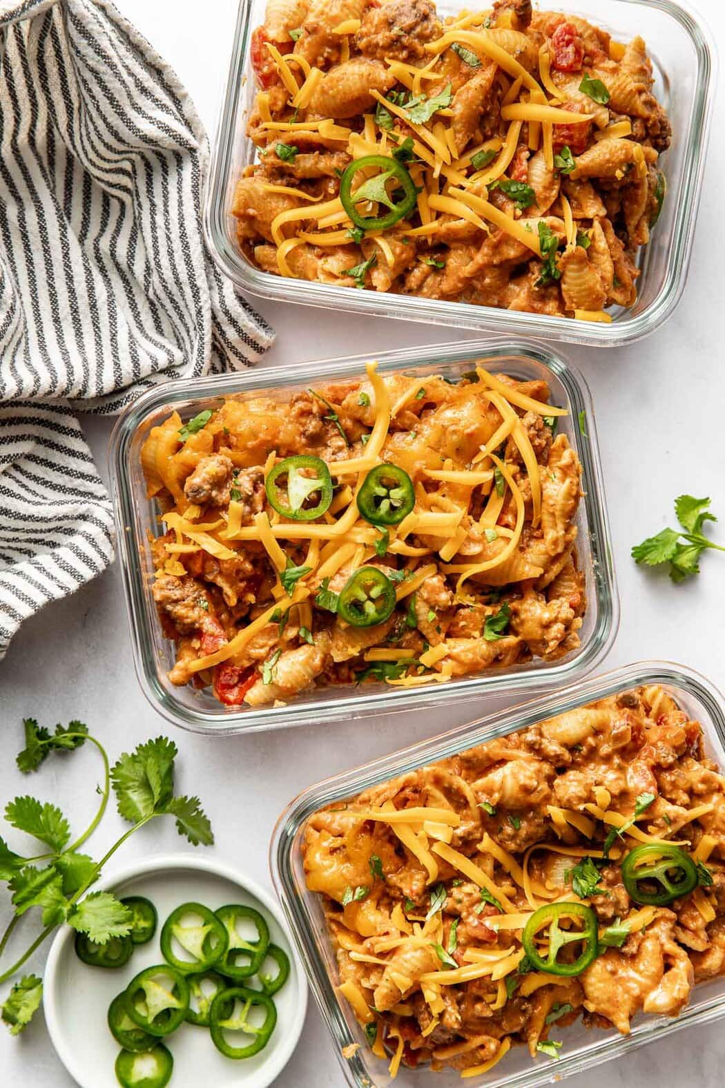 Overhead view of three glass meal prep containers filled with Taco Pasta and topped with sliced jalapenos and sprinkled with fresh cilantro.