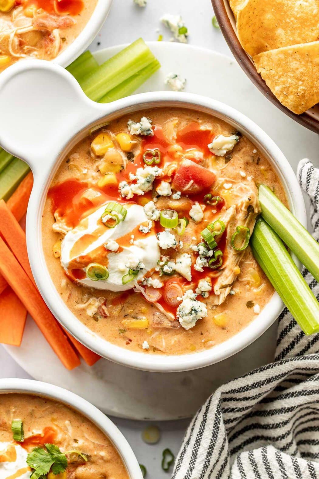 Close up view of a bowl of Buffalo Chicken Chili with two celery sticks on the side topped with bleu cheese crumbles and drizzled with buffalo sauce.