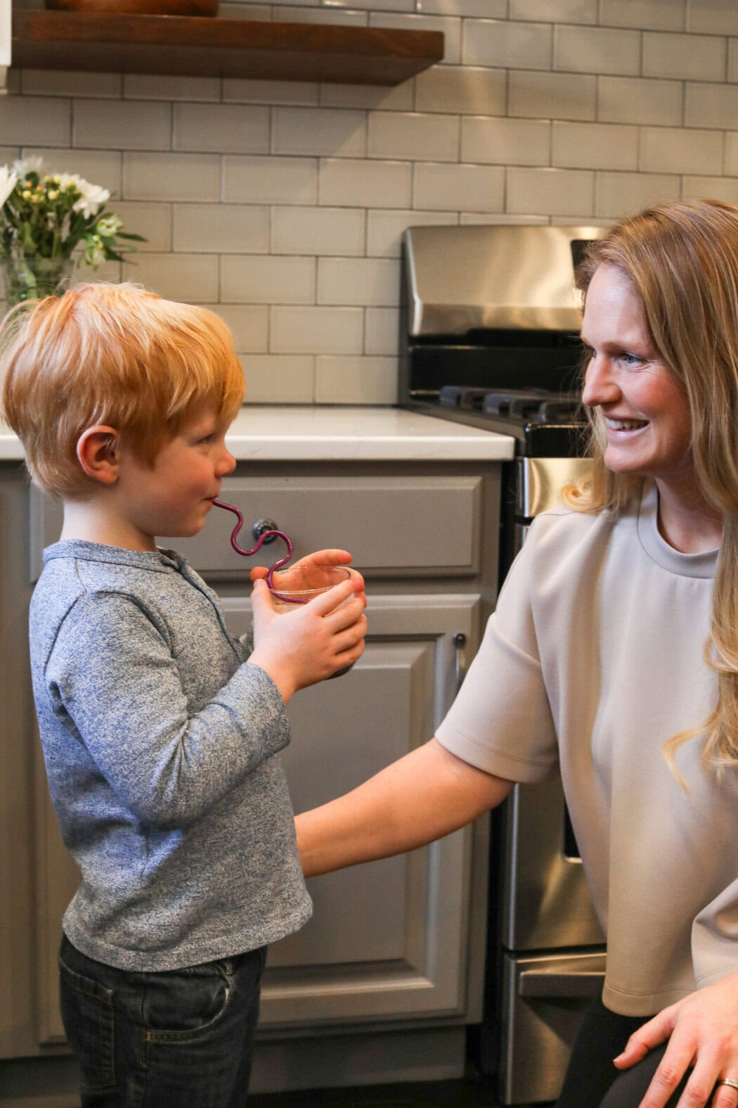 A mother and son in a kitchen smiling at each other while the little boy sips a hiya drink with a fun straw.